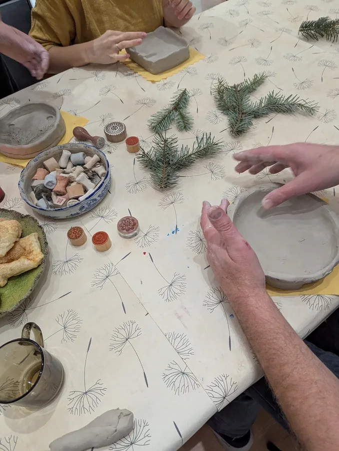 Praca z gliną przy stole podczas dodatkowego doszkolenia SensoCeramiki; obok widoczne są gałązki iglaste, stemple i materiały do pracy.